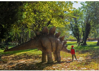 The image shows a boy standing next to a Stegosaurus dinosaur in a park.