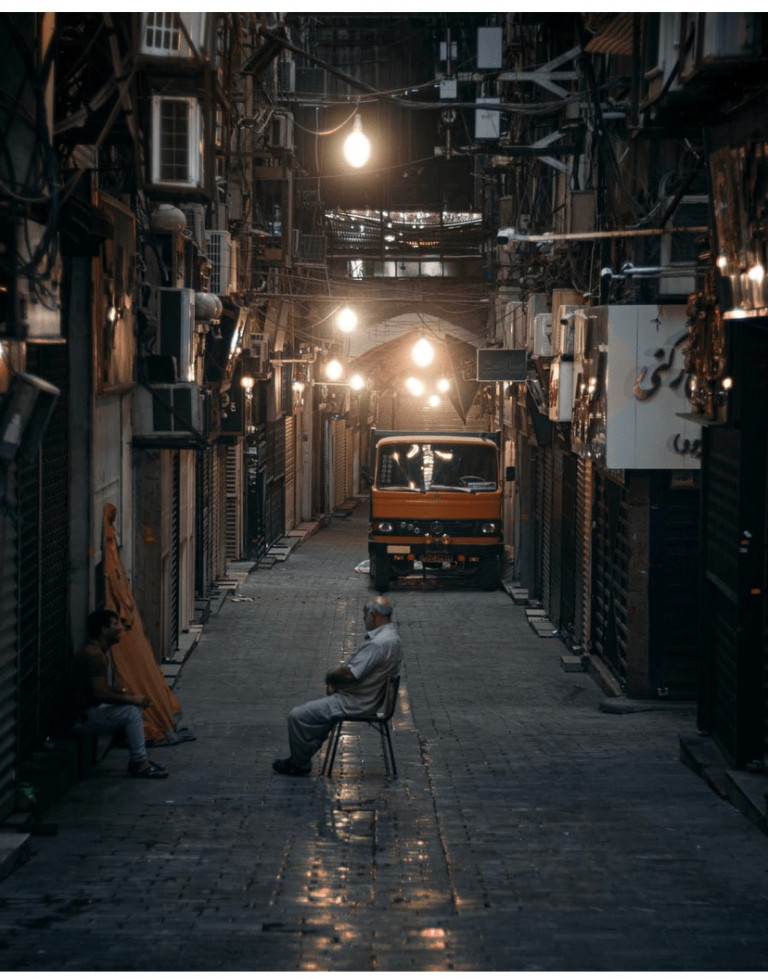 The image shows a covered paved street. One person is sitting in a chair in the middle of the street talking to another sitting at the side walk. There is a truck parked in the street behind them. The street is lighted up well with lights hanging from the ceiling.