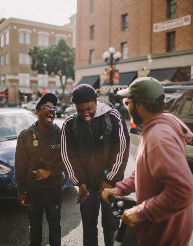 The image shows three young men standing by the roadside and laughing at a shared joke.