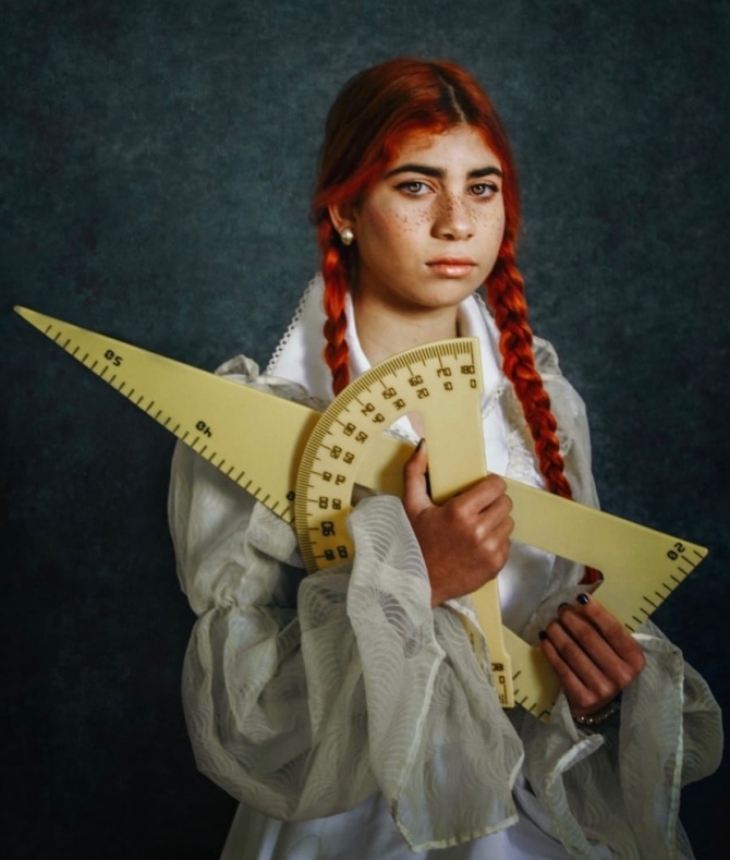 The image shows a girl carrying an oversized geometry set in her arms, looking directly at the camera.
